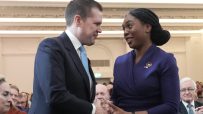 Kemi Badenoch and Robert Jenrick shaking hands at an event, with attendees clapping in the background.