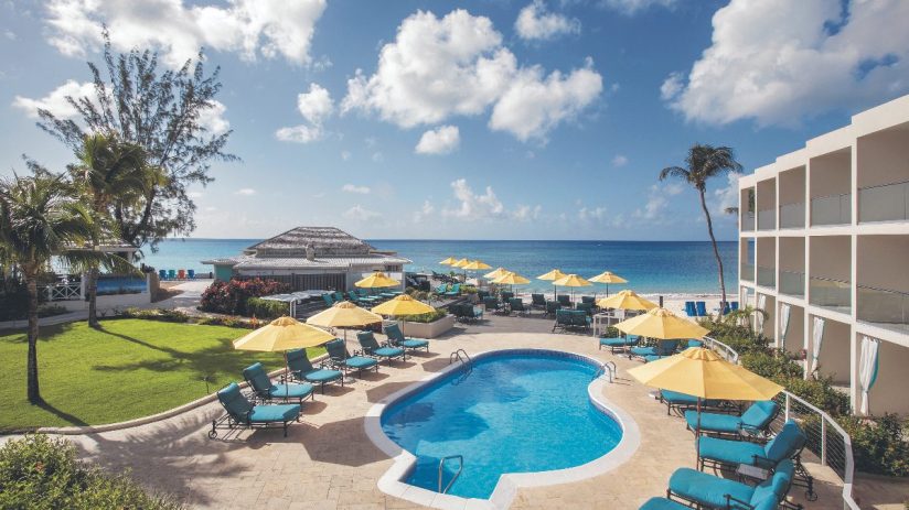 Resort pool and lounge chairs overlooking a Barbados beach with palm trees and a hotel building under a blue sky.