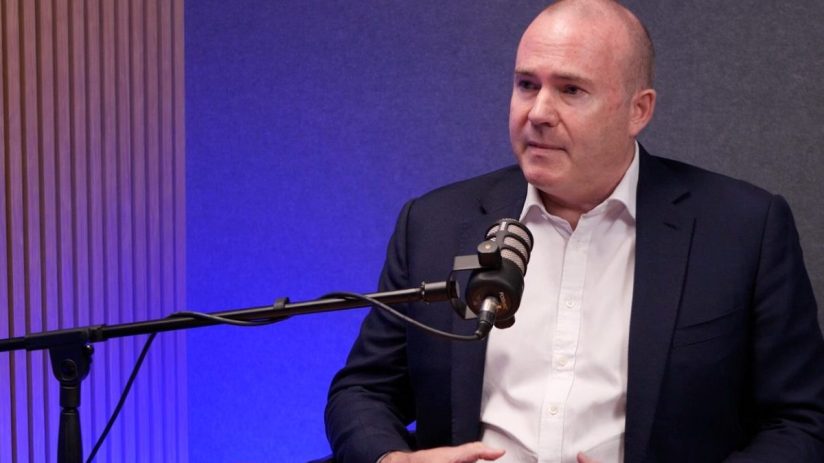 Bald man in suit at Barings, speaking into a podcast microphone in a studio with blue lighting.