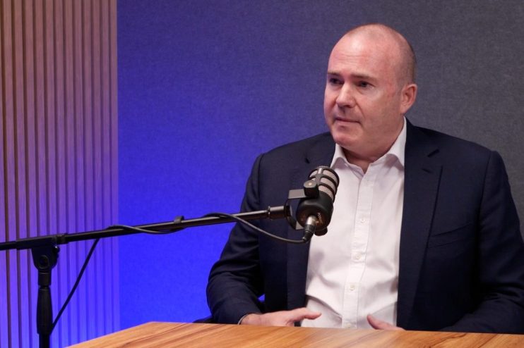 Bald man in suit at Barings, speaking into a podcast microphone in a studio with blue lighting.