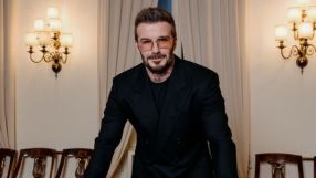 David Beckham in a dark suit and amber-tinted glasses, standing in a room with ornate wall sconces.
