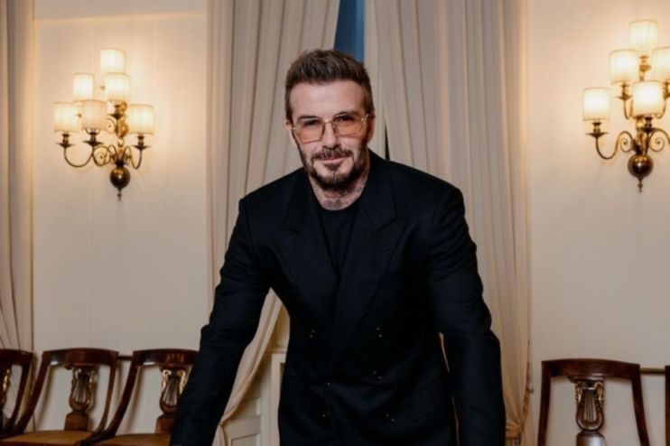 David Beckham in a dark suit and amber-tinted glasses, standing in a room with ornate wall sconces.