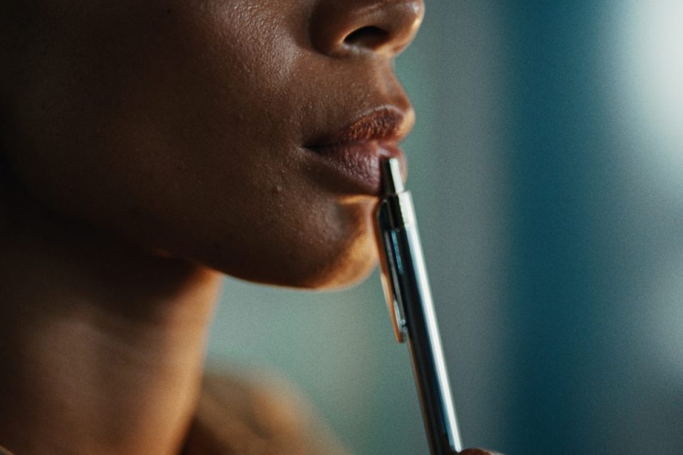 Close-up of a persons mouth with a silver pen held to their lower lip, implying thought or concentration