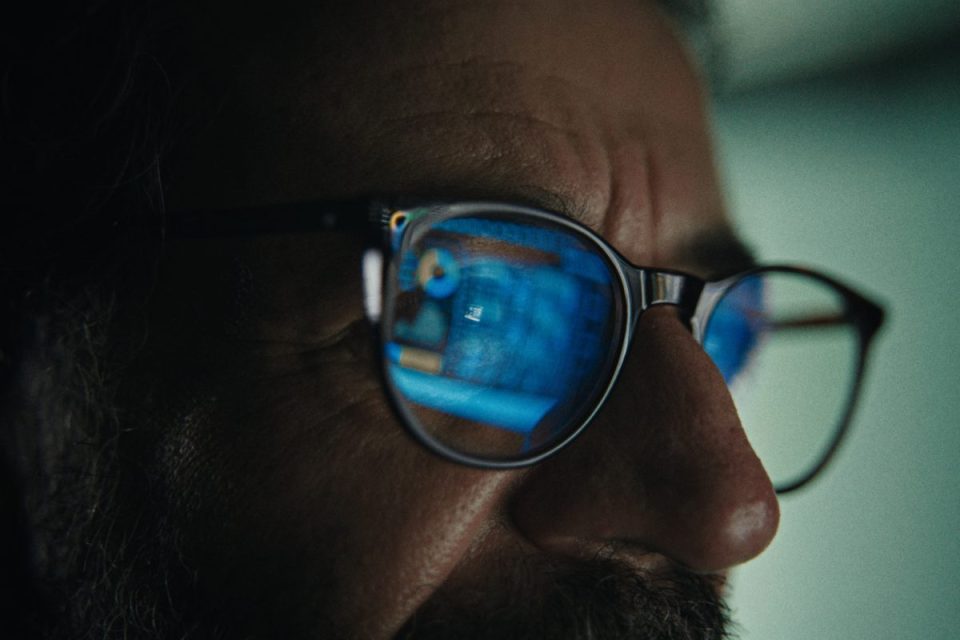 Close-up of a persons face with glasses reflecting a blue computer screen, indicating digital work or data analysis.
