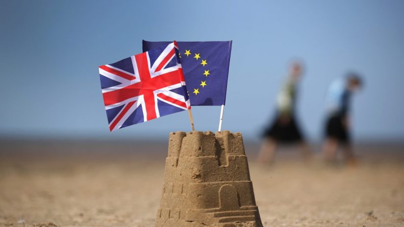 UK and EU flags planted in a sandcastle on a beach, symbolizing Brexit negotiations.