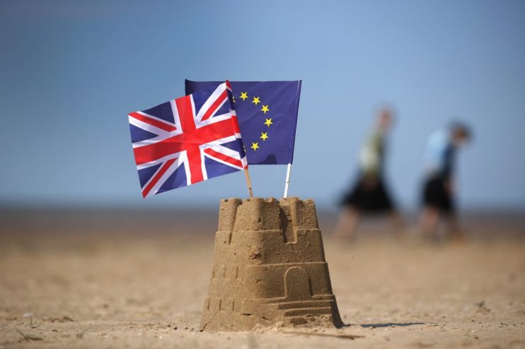 UK and EU flags planted in a sandcastle on a beach, symbolizing Brexit negotiations.