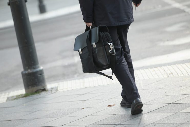Man in dark suit carrying a black leather briefcase, walking on a city sidewalk.