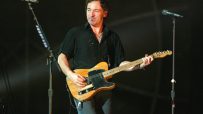 Bruce Springsteen performing live, playing an electric guitar, with microphones on stands in a dark background