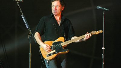 Bruce Springsteen performing live, playing an electric guitar, with microphones on stands in a dark background