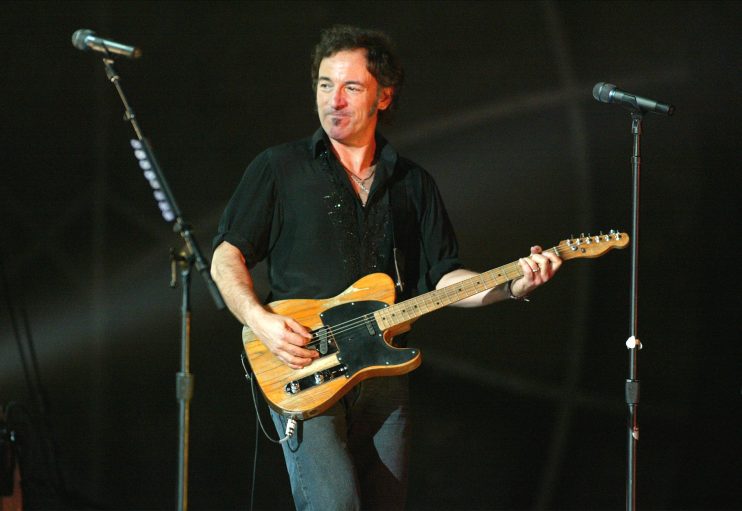 Bruce Springsteen performing live, playing an electric guitar, with microphones on stands in a dark background