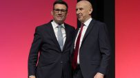 Andy Burnham and Jon Healey smiling in dark suits against a red background