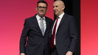 Andy Burnham and Jon Healey smiling in dark suits against a red background