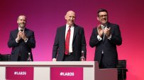 Andy Burnham, Peter Kyle, and Jonathan Reynolds applauding at a #LAB26 event with a pink background.