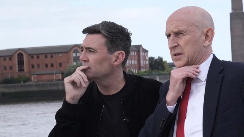 Andy Burnham and Paul Healey on a boat, looking out over water with buildings in the background.