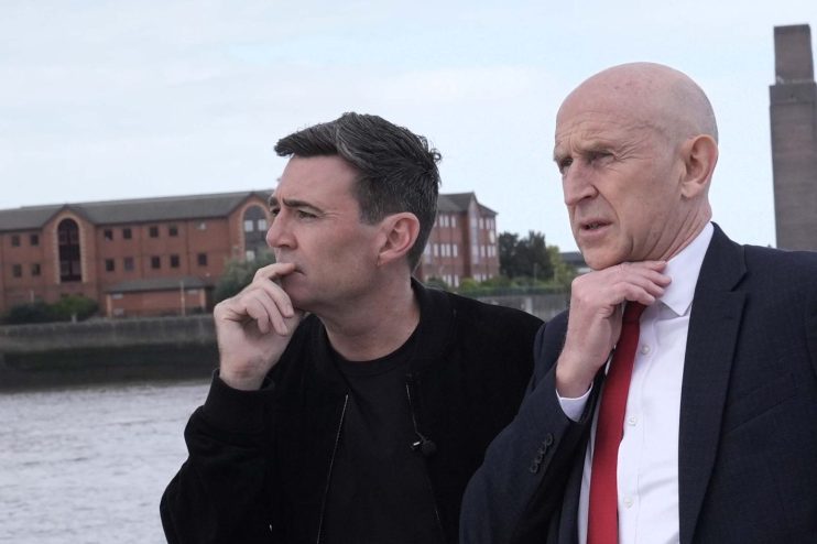 Andy Burnham and Paul Healey on a boat, looking out over water with buildings in the background.