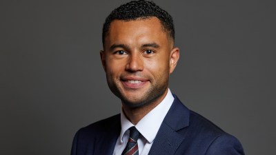 Callum Anderson, a smiling man in a navy suit and patterned tie, against a dark gray background.