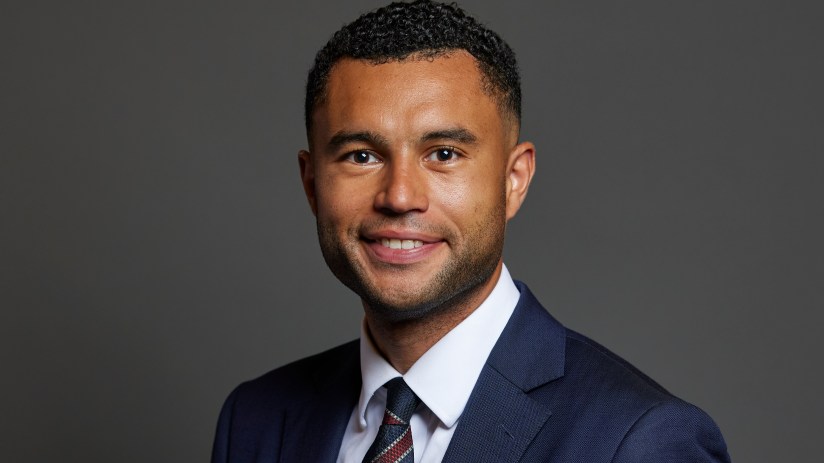 Callum Anderson, a smiling man in a navy suit and patterned tie, against a dark gray background.