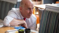 Chancellor John Healey in a white shirt and red tie, leaning on a table with plates of food in a restaurant