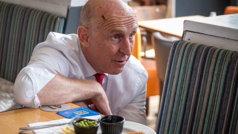 Chancellor John Healey in a white shirt and red tie, leaning on a table with plates of food in a restaurant