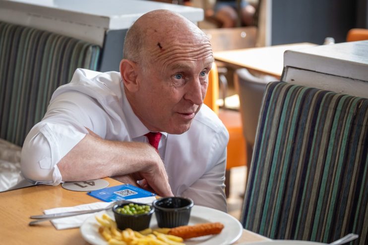 Chancellor John Healey in a white shirt and red tie, leaning on a table with plates of food in a restaurant