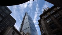 Glass skyscraper rising into a cloudy blue sky, surrounded by dark historic London buildings.
