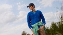 Man in blue Crew Clothing cotton cable knit cricket jumper, khaki trousers, and dark cap outdoors.