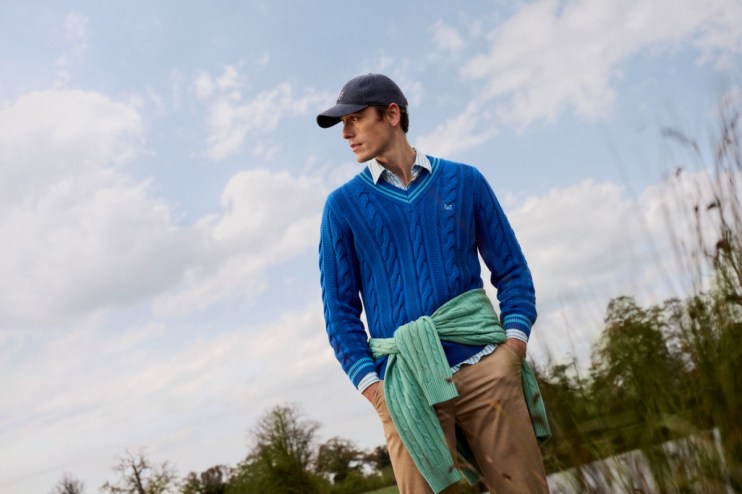Man in blue Crew Clothing cotton cable knit cricket jumper, khaki trousers, and dark cap outdoors.