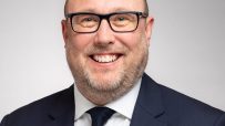 David Bimpson, smiling professional in a navy suit and glasses, white shirt, dark knitted tie