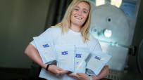 Smiling blonde woman in white shirt holding four pouches of Ruby Alba products in an industrial setting