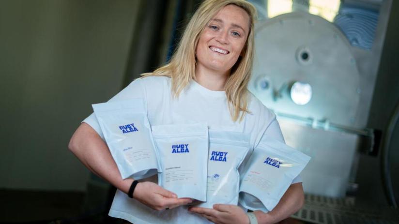 Smiling blonde woman in white shirt holding four pouches of Ruby Alba products in an industrial setting