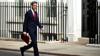 Ed Miliband, in a suit and red tie, strides past Downing Street railings with a red folder in hand.