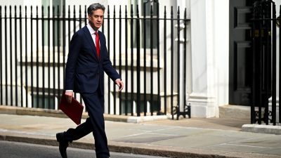 Ed Miliband in a suit and red tie, carrying a red folder, walking past railings outside Downing Street