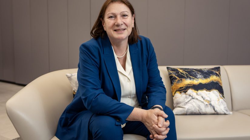 Professional woman in a blue suit and pearl necklace, smiling while seated on a light-colored sofa.