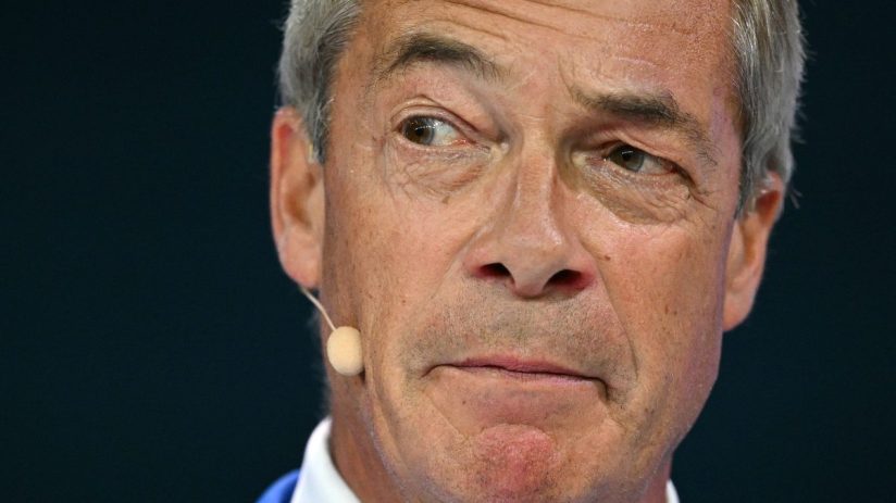 Nigel Farage close-up, looking left, with a microphone near his ear, against a dark background