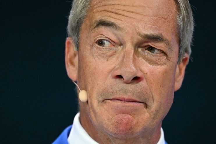 Nigel Farage close-up, looking left, with a microphone near his ear, against a dark background