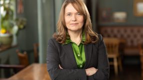 Fiona Dickie, a woman in a dark pinstripe suit and green shirt, standing with arms crossed in an office