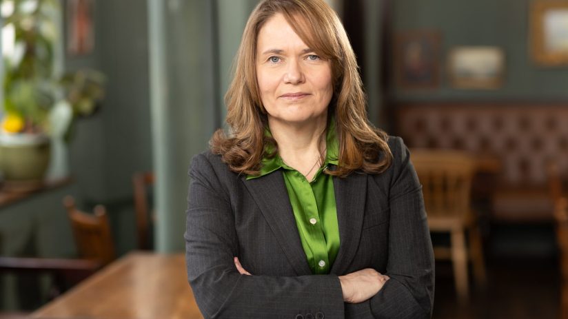 Fiona Dickie, a woman in a dark pinstripe suit and green shirt, standing with arms crossed in an office