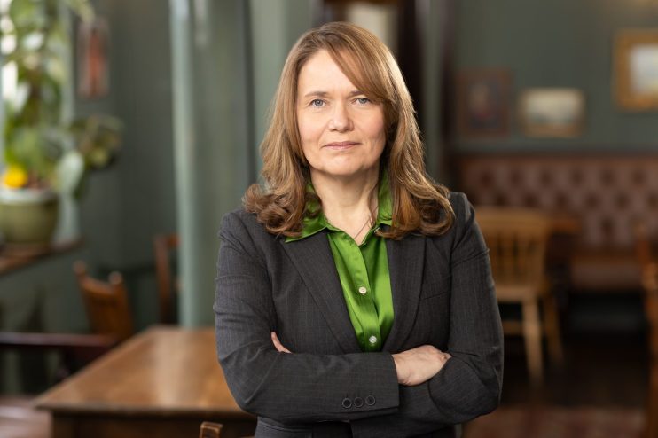 Fiona Dickie, a woman in a dark pinstripe suit and green shirt, standing with arms crossed in an office