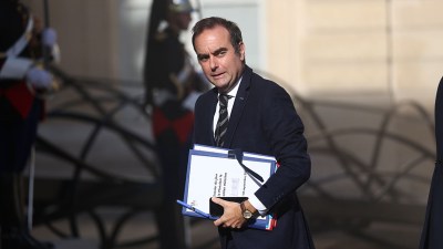 S&eacute;bastien Lecornu, French Prime Minister, carries documents and a phone, with uniformed guards in the background.