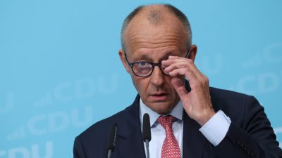 Friedrich Merz in a suit and red tie, adjusting his glasses at a podium with microphones, against a blue background.