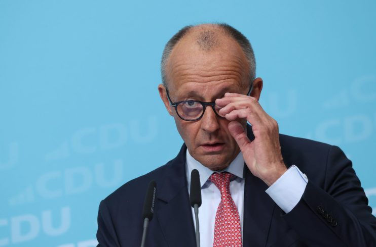 Friedrich Merz in a suit and red tie, adjusting his glasses at a podium with microphones, against a blue background.