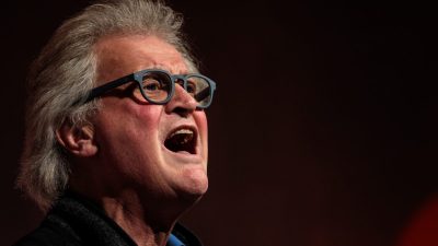 Older man with grey hair and blue glasses speaking passionately, mouth open, against a dark background.