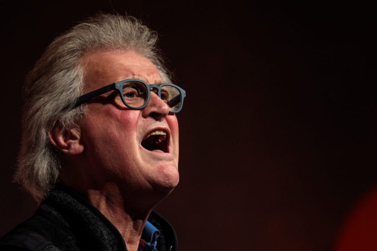 Older man with grey hair and blue glasses speaking passionately, mouth open, against a dark background.