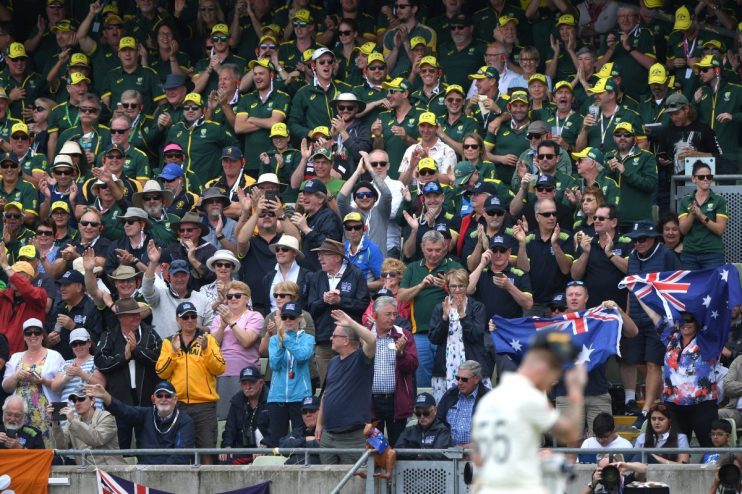 Crowd of Australian cricket fans cheering, many wearing green and gold uniforms and hats, some holding flags.