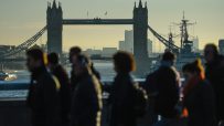 Silhouetted pedestrians walk on a bridge with Londons Tower Bridge and the River Thames in the background.