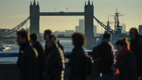 Silhouetted pedestrians walk on a bridge with Londons Tower Bridge and the River Thames in the background.