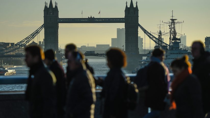 Silhouetted pedestrians walk on a bridge with Londons Tower Bridge and the River Thames in the background.
