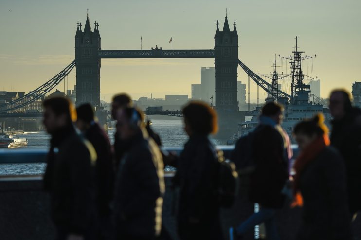 Silhouetted pedestrians walk on a bridge with Londons Tower Bridge and the River Thames in the background.