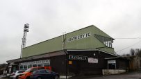 Exterior view of Bath City FC stadium with Randalls signage, parked cars, and a tall floodlight against a cloudy sky.
