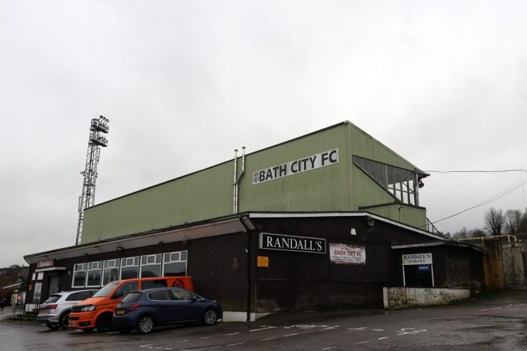 Exterior view of Bath City FC stadium with Randalls signage, parked cars, and a tall floodlight against a cloudy sky.